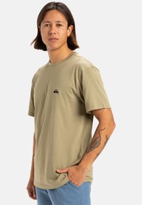 Camiseta de manga corta verde oliva en tejido suave con cuello redondo y un pequeño logo negro en el lado izquierdo del pecho. Ajuste casual, textura lisa.