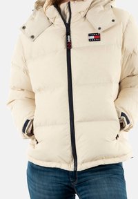 Beige vadderad jacka med huva, framhävd av en svart dragkedja, sidofickor och en liten logotyp. Mjuk, quiltad textur.