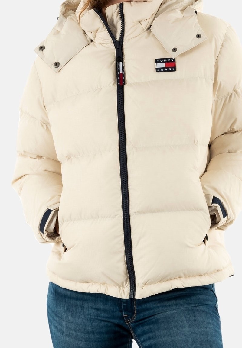 Beige vadderad jacka med huva, framhävd av en svart dragkedja, sidofickor och en liten logotyp. Mjuk, quiltad textur.