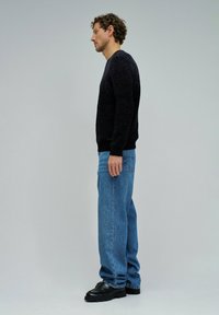 Un uomo in maglione nero, jeans blu e scarpe nere in profilo contro uno sfondo grigio chiaro uniforme.