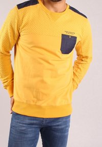Felpa gialla con maniche testurizzate e una tasca blu scuro. Presenta un collo a V e polsini a costine, evidenziando un design casual.