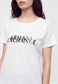 Γυναίκα που φοράει ένα λευκό t-shirt με μαύρο και χρυσό πολυεπίπεδο κείμενο "ARMANI EXCHANGE" στο στήθος, στέκεται σε απλό φόντο.