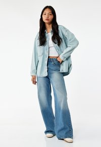 Giacca di jeans oversize azzurro chiaro, canotta bianca aderente, jeans a vita alta svasati e scarpe beige su uno sfondo neutro.