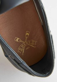 Tumed sinine nahast jalanõu sisemus, millel on kuldne logo ristatud peatükkidega, sile pruun nahast vooder ja detailne tikand servade ääres.