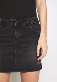 Jupe en jean noire avec bouton devant et poches, portée avec un haut noir, montrant le bras droit et la main reposant à côté de la jupe.