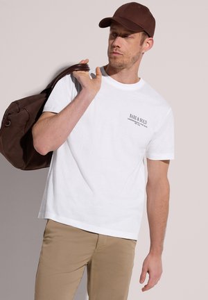 Homme portant une casquette marron, un t-shirt blanc « Bare & Bold » et un pantalon beige, portant un sac de sport en cuir marron sur l'épaule.