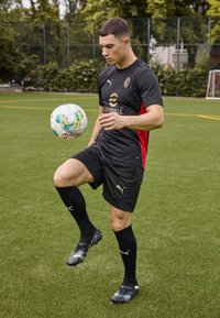 Svart och röd fotbollskit med logotyp, svarta strumpor och svarta skor. Spelaren balanserar en vit fotboll på ett knä på konstgräs.
