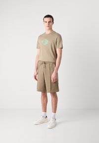 Beige T-skjorte med en grønn diamantlogo, parret med beige shorts med sidelommer. Modellen bruker hvite joggesko og ankelsokker.