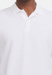 Weiße Poloshirt aus strukturiertem Stoff mit klassischem Kragen, Knopfblende und dezentem Logo auf der Brust.