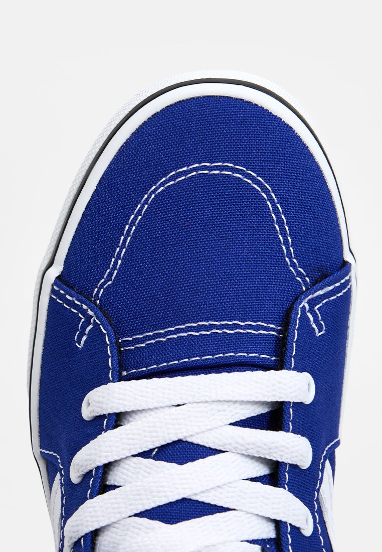 Zapatilla de lona azul con cordones blancos, suela de goma blanca y detalles de costura en contraste en la parte superior. Diseño de punta redondeada.
