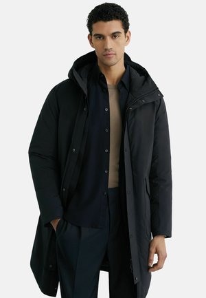 Mężczyzna w długim czarnym pikowanym płaszczu, pod którym ma czarną koszulę na guziki i beżową koszulkę, stoi z jedną ręką w kieszeni.