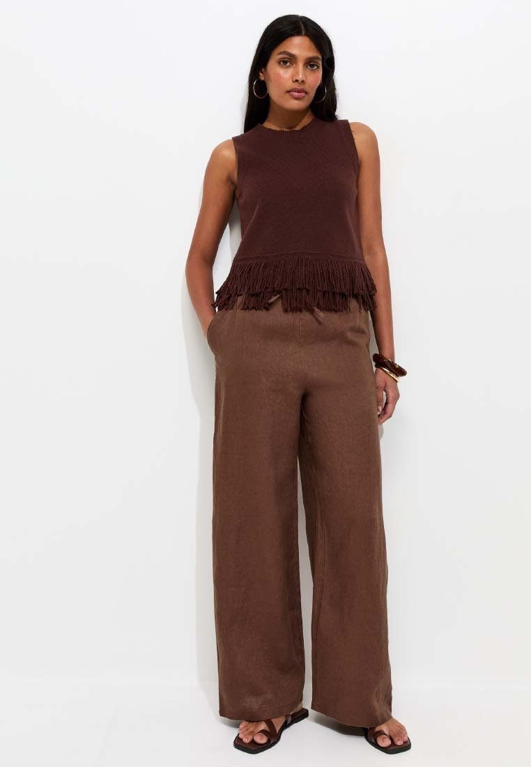Woman with long dark hair wearing sleeveless brown fringed top, wide-leg brown pants, black sandals, and hoop earrings standing against white wall.