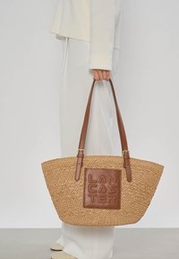 Main tenant un grand sac fourre-tout en paille tressée avec des poignées en cuir marron et un patch carré embossé des lettres "LANCASTER".