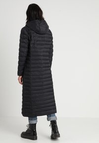 Abrigo puffer negro largo con un patrón de acolchado horizontal, capucha y silueta recta. Combinado con jeans acampanados y botas negras con cordones.