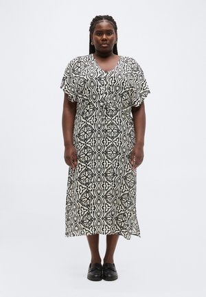 Kvinde med fletninger iført en sort og hvid mønstret midikjole og sorte slip-on sko, stående foran en ensfarvet hvid baggrund.