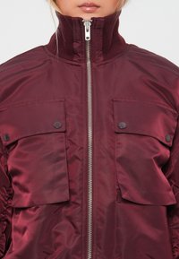 Burgundy insulated jacket with a high collar, two front flap pockets, and a full-length zipper. Smooth, shiny fabric with a textured finish.