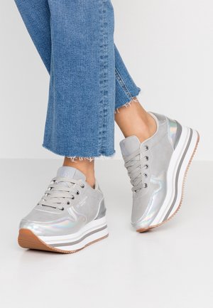 Stopy noszące szare sneakersy na platformie z iryzującymi detalami i białymi podeszwami, zestawione z krótkimi niebieskimi dżinsami.