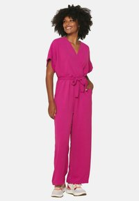 Fuchsia jumpsuit with short sleeves, wrap-style neckline, tie waist, and wide leg design. Paired with sneakers, showcasing a casual look.