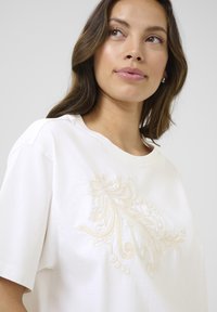 T-shirt en coton blanc avec des broderies crémeuses intriquées dans un motif floral sur le devant. Coupe décontractée avec des manches courtes et un col rond.