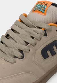 Beżowe zamszowe sneakersy z fakturowanymi jasnoszarymi sznurówkami, granatowymi akcentami i pomarańczową etykietą na języku. Posiadają wyściełany kołnierz dla wygody.