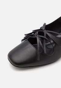 Bailarinas de cuero negro con punta redonda, que presentan un detalle de cordones entrelazados en la parte delantera y una superficie suave y texturizada.