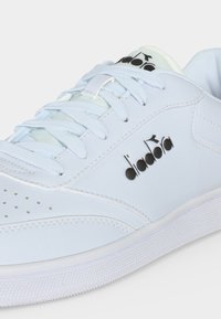 Λευκό αθλητικό παπούτσι Diadora με λευκά κορδόνια, διάτρητη μύτη και μαύρο λογότυπο Diadora στο πλάι και στη γλώσσα.