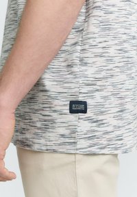 Chemise à manches courtes, de couleur claire, avec un motif abstrait texturé dans des tons de gris, rose et blanc. Présente une étiquette de marque à l'ourlet.