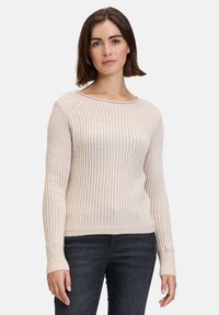 Beige gestrickter Pullover mit langen Ärmeln, geripptem Muster, weitem Ausschnitt und enganliegenden Bündchen, kombiniert mit dunklen Denim-Jeans.