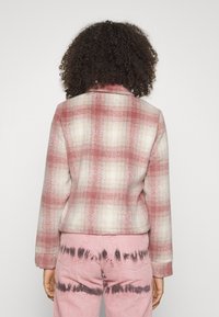 ternet jakke i lyserød og creme med krave og en kasseformet pasform. Kombination med lyserøde jeans, der har et mørkt batikmønster på benene.