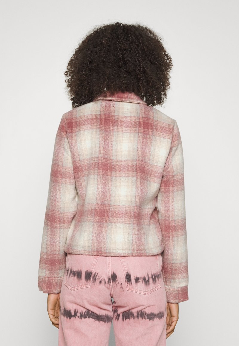 ternet jakke i lyserød og creme med krave og en kasseformet pasform. Kombination med lyserøde jeans, der har et mørkt batikmønster på benene.