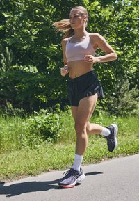 Femme coureuse portant un soutien-gorge de sport rose clair et un short noir, avec des chaussures de course noires à semelles blanches et des chaussettes blanches, sur un chemin entouré de verdure.