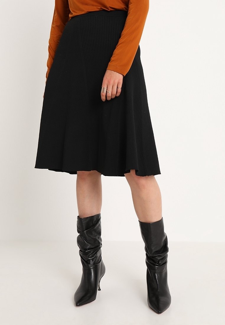 Femme portant une jupe noire évasée longueur genou, des bottes à talons noires et amples, et un haut marron à manches longues, debout devant un fond uni.