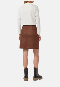 Brązowa zamszowa spódnica z widocznymi szwami, w połączeniu z białą koszulą z długim rękawem oraz czarnymi botkami na grubych podeszwach z elastycznymi panelami.
