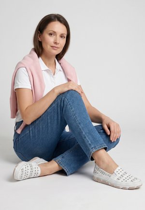 Vrouw zittend met gekruiste benen, draag een wit poloshirt, een roze trui over de schouders gedrapeerd, blauwe spijkerbroek en witte geperforeerde slip-on schoenen.