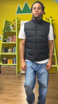 Crna podstavljena prsluk preko bijele majice kratkih rukava, u kombinaciji sa svijetloplavim trapericama. U pozadini žuti zidovi i zelene sobne biljke.