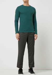 Dunkeltealfarbenes Langarmshirt, leicht und körpernah geschnitten, kombiniert mit grauen, zulaufenden Hosen und schwarzen Turnschuhen. Minimalistisches Design, ohne Muster.