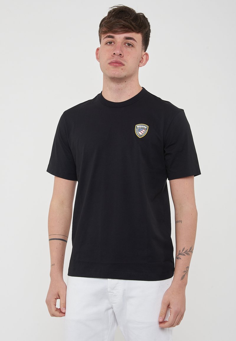 Maglietta nera in cotone con maniche corte, scollatura rotonda e un logo sulla parte sinistra del petto. Design semplice, senza motivi o accenti visibili.