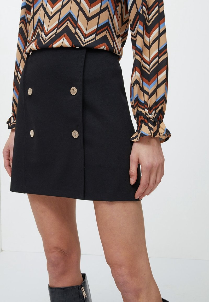 Černá mini sukně s dvojicí zlatých knoflíků vpředu, s přiléhavým střihem a hladkou texturou, doplněná vzorovanou blůzou.