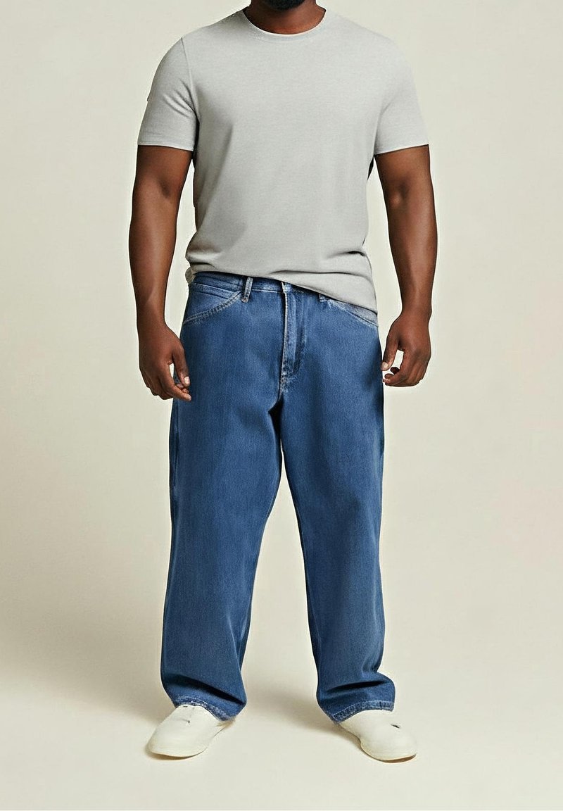Homme portant un t-shirt gris clair à manches courtes, un jean bleu ample et des baskets blanches, debout devant un fond uni.