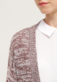 Cardigan lavorato a maglia marrone e bianco con apertura frontale, caratterizzato da un motivo testurizzato e maniche larghe. Indossato sopra una camicia bianca con colletto.