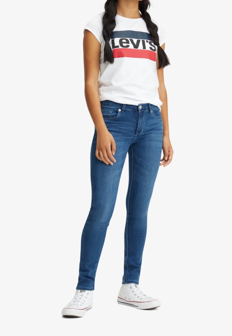 Femme portant un t-shirt blanc Levi's, un jean skinny bleu et des baskets montantes blanches, debout, les bras détendus le long du corps.