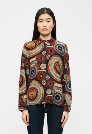 Femme portant une chemise à manches longues boutonnée avec des motifs circulaires multicolores et abstraits, se tenant devant un fond blanc uni.