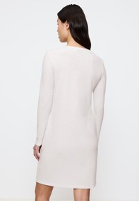 Donna con lunghi capelli scuri indossa un vestito in maglia leggero beige, a maniche lunghe e lunghezza al ginocchio, in piedi di spalle contro uno sfondo bianco semplice.