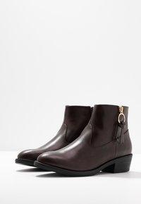 Botas de cuero marrón hasta el tobillo con una textura suave, punta redondeada, tacón bajo y cuadrado, y acento decorativo de borlas cerca de la cremallera.