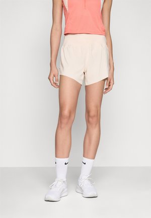 Lette beige treningsshorts med en tettsittende midje, skrått skjær og sidelommer, kombinert med en korallfarget ermeløs topp og hvite joggesko.