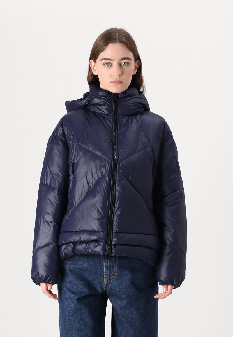 Navy pufīgs mētelis ar augstu apkakli, rāvējslēdzēja aizdari un sānu kabatām, kas raksturojas ar quilted zīmējumu. Gluda, spīdīga auduma struktūra ar brīvu piegriezumu.