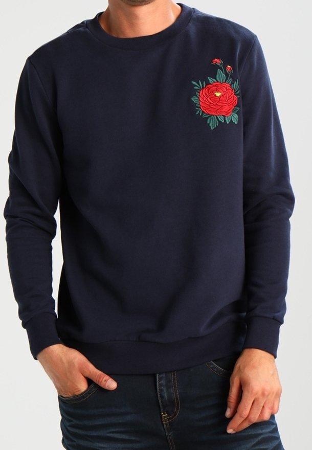 Man draagt een marineblauwe trui met een rode bloemenborduur op de linkerborst, handen in de zakken, donkere spijkerbroek zichtbaar.
