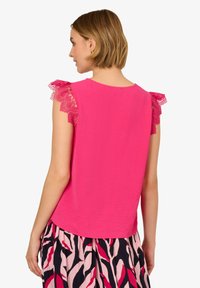Top senza maniche rosa con dettagli in pizzo sulle spalle, texture morbida, scollatura rotonda e orlo leggermente curvato, indossato con una gonna a fantasia.