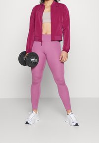 Mujer en mallas y chaqueta de entrenamiento magenta sosteniendo un dumbbell negro, usando zapatillas blancas, de pie contra un fondo liso.