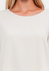 Ανοιχτό μπεζ τοπ με απαλή υφή, διαθέτει πλατιά λαιμόκοψη και χαλαρή γραμμή, φτιαγμένο από μαλακό, ροή ύφασμα.